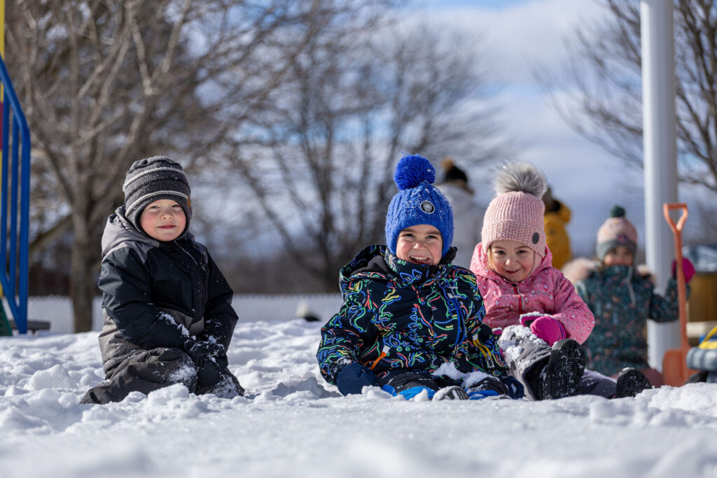 Photo d'un groupe d'enfants souriants du CPE Flocon de rêve, assis dans la neige, à l'extérieur en hiver