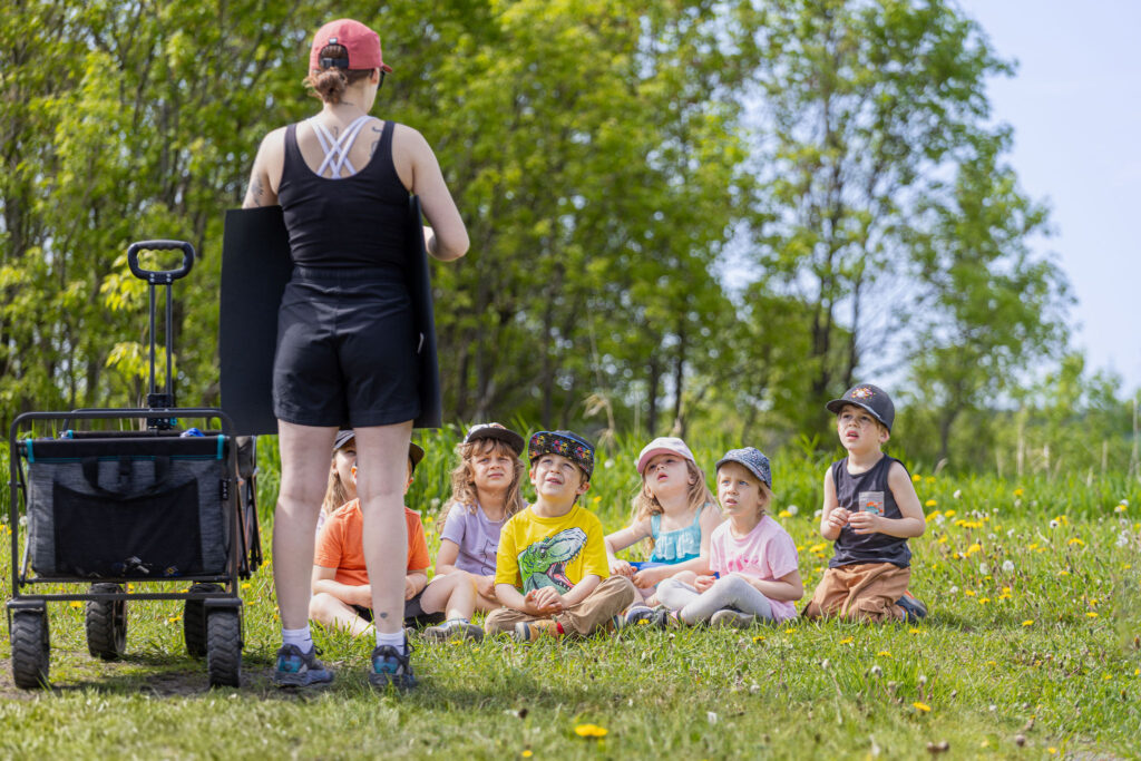 Photo d'un groupe d'enfants du CPE Flocon de rêve, assis dans l'herbe à l'extérieur en été, supervisé par une éducatrice