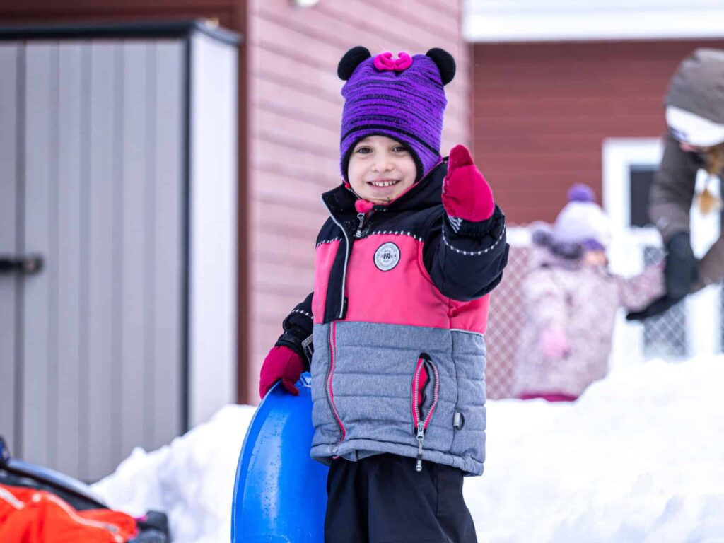 Photo d'une petite fille du CPE Flocon de rêve dehors en hiver, tenant une luge et souriant, pouce vers le haut